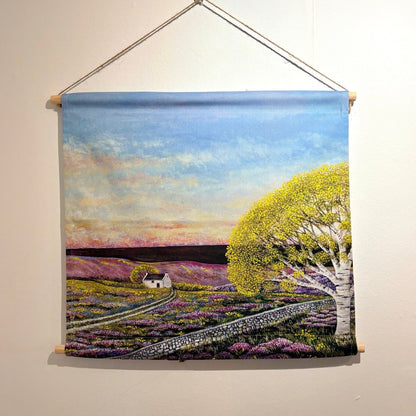 A woven wall hanging tapestry depicting a rural landscape with a blue sky, purple flower fields, a small white cottage, and a large yellow-green tree, suspended from a wooden rod against a plain wall.