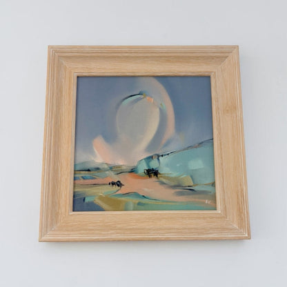 A framed abstract landscape painting featuring soft brushstrokes of blue, teal, peach, and beige, housed in a light natural oak wood frame against a plain white wall.