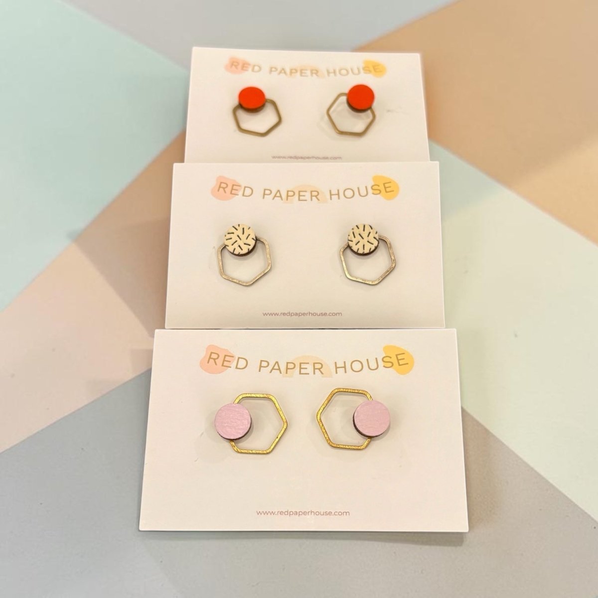 Three pairs of geometric stud earrings displayed on white cards. The top pair features red-orange circular studs with gold hexagonal wire frames; the middle pair has gold textured circular studs with hexagonal wire frames; the bottom pair shows light pink circular studs with matching hexagonal wire frames.