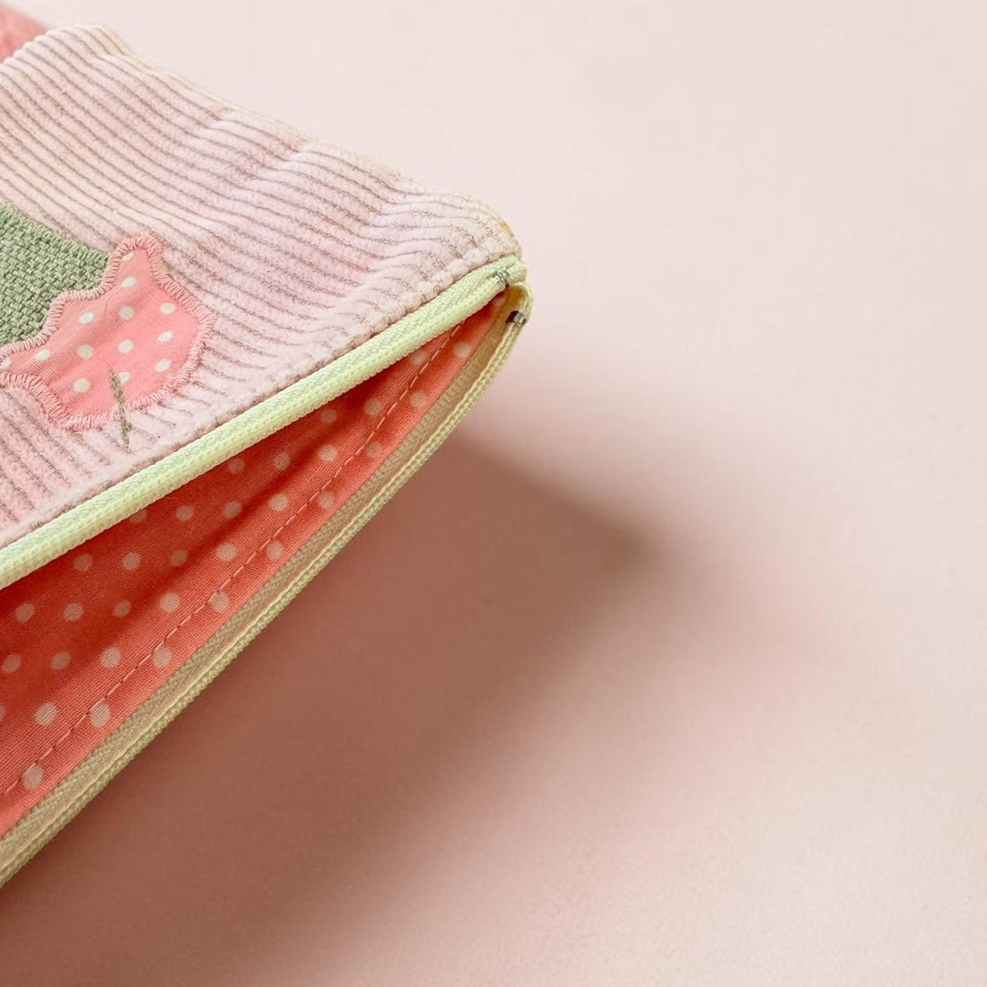 A close-up of a pastel pink zippered pouch with a ribbed fabric exterior, a green and pink applique detail, and a coral polka-dot interior lining, set against a solid pale pink background.