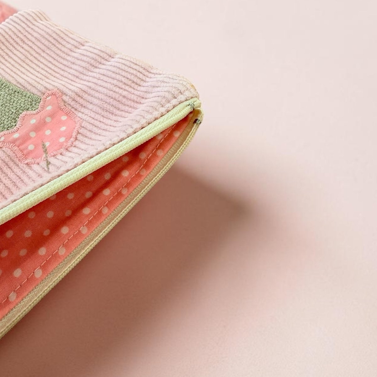 A close-up of a pastel pink zippered pouch with a ribbed fabric exterior, a green and pink applique detail, and a coral polka-dot interior lining, set against a solid pale pink background.