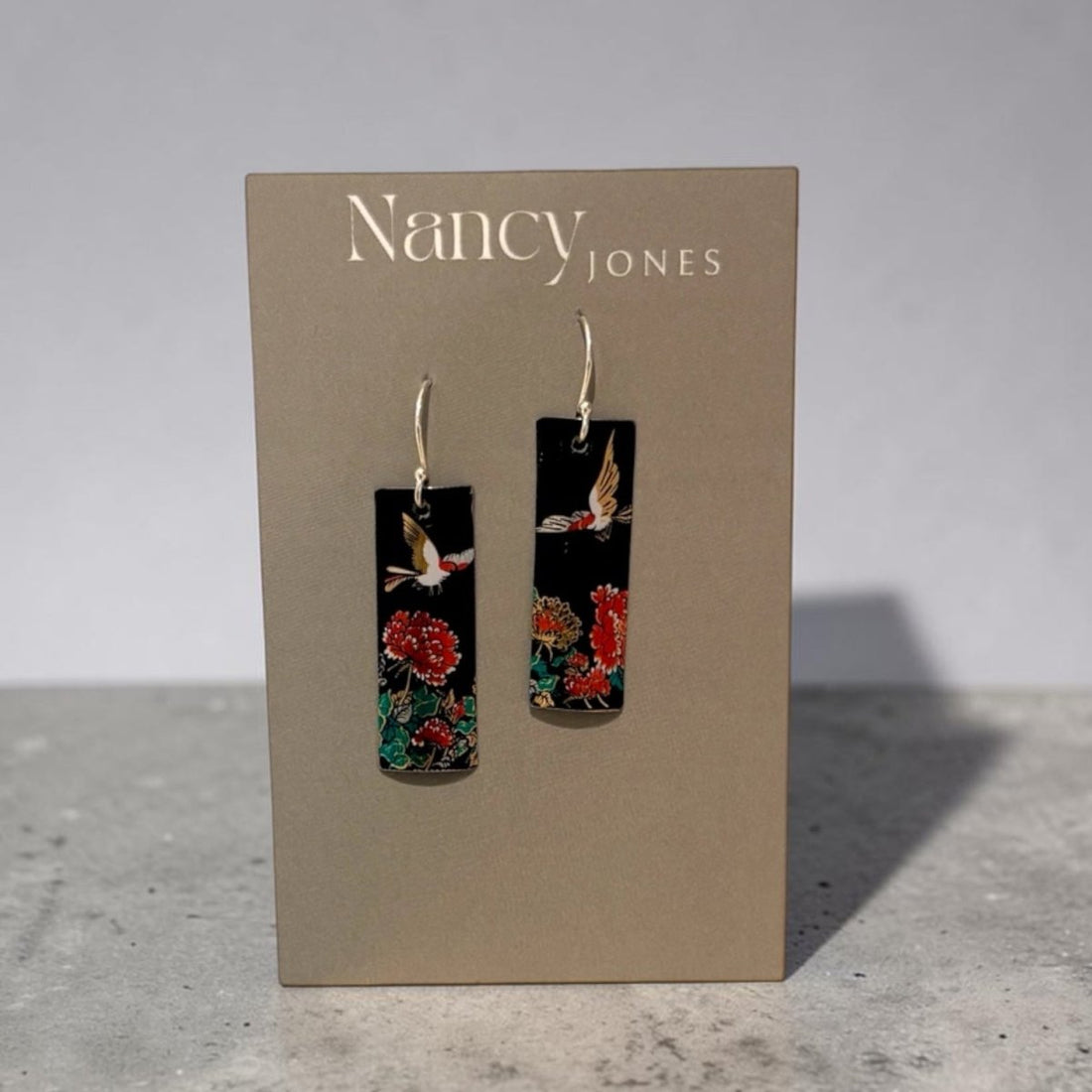 A pair of Nancy Jones dangle earrings displayed on a taupe jewelry card. The earrings feature rectangular black pendants decorated with red and orange flowers and white birds, suspended from silver hooks.