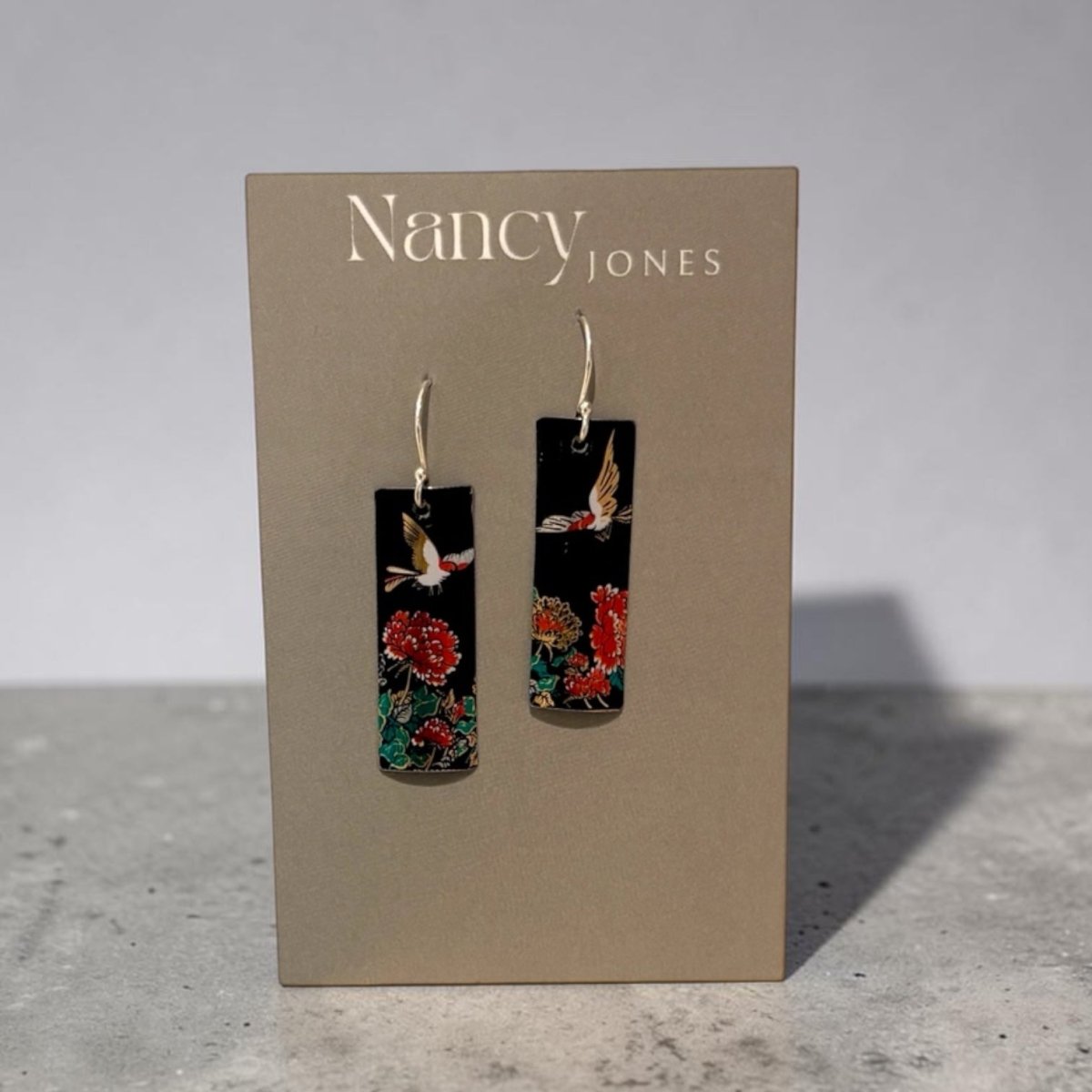 A pair of Nancy Jones dangle earrings displayed on a taupe jewelry card. The earrings feature rectangular black pendants decorated with red and orange flowers and white birds, suspended from silver hooks.