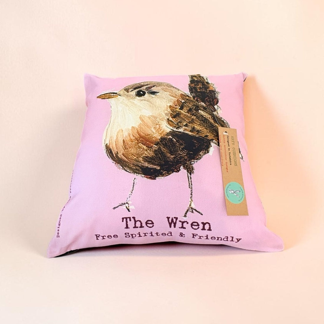 A square pink pillow featuring a painted bird illustration labeled &