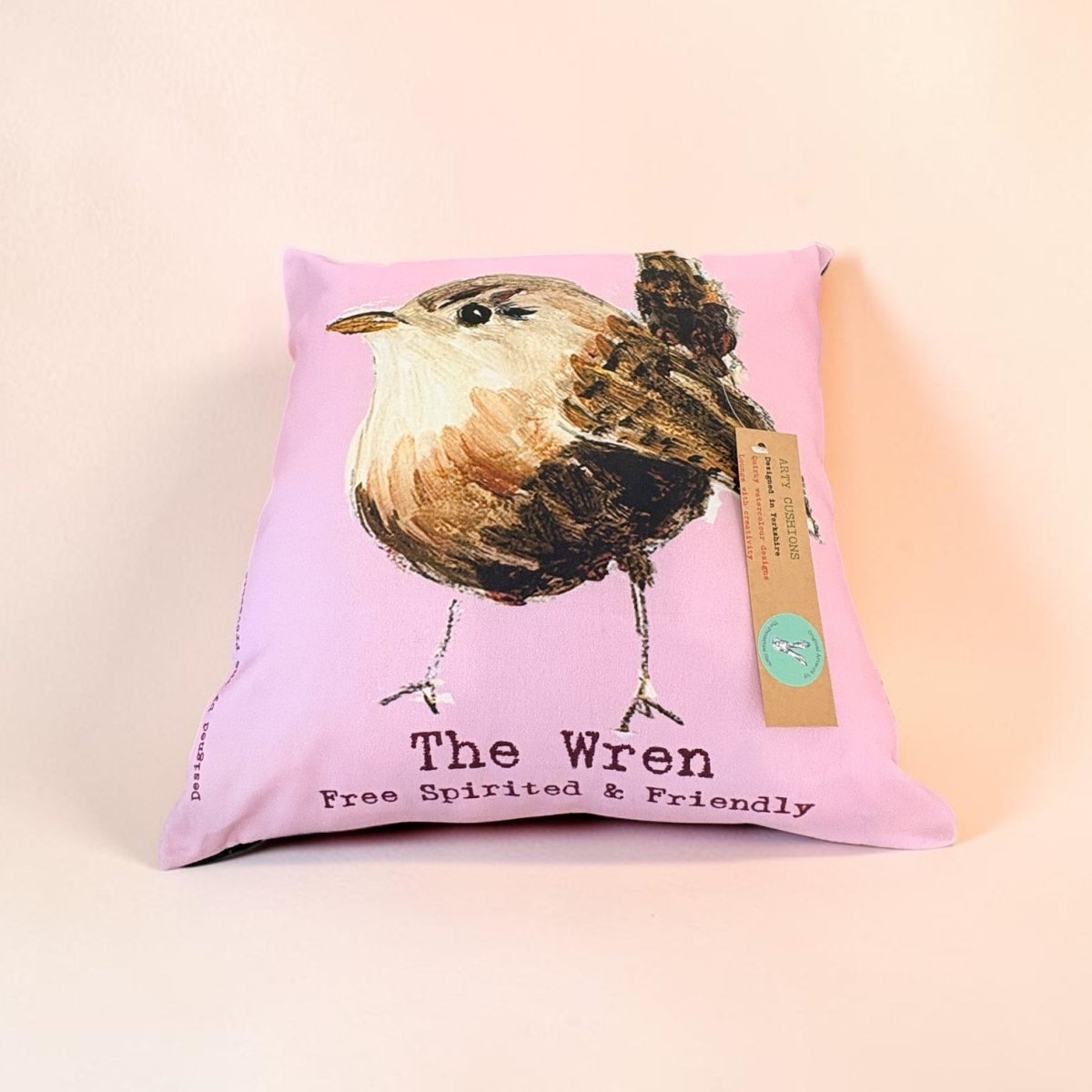 A square pink pillow featuring a painted bird illustration labeled &