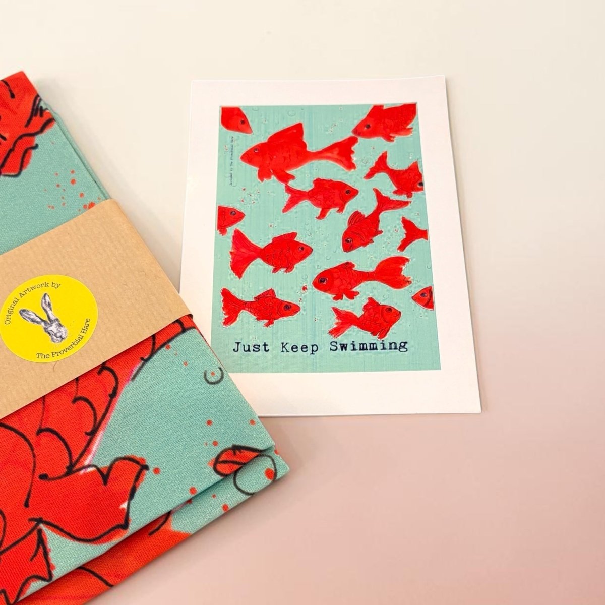 A folded fabric art print or card with a turquoise background and red fish pattern, placed beside a matching folded textile wrapped with a kraft paper band and a round yellow sticker. The printed card reads &