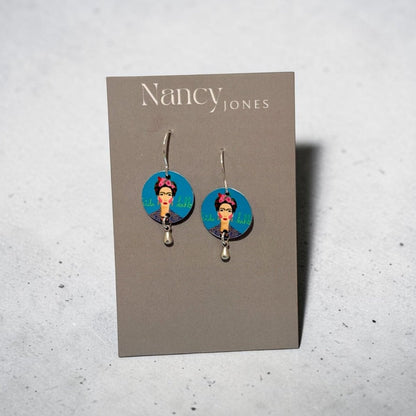 A pair of Nancy Jones dangle earrings displayed on a gray card. The earrings feature circular blue and pink graphic charms with a portrait illustration, suspended from silver hooks against a light gray textured background.