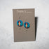A pair of Nancy Jones dangle earrings displayed on a gray card. The earrings feature circular blue and pink graphic charms with a portrait illustration, suspended from silver hooks against a light gray textured background.