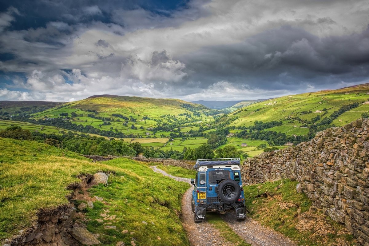 Blue off-road vehicle on a dirt path with green hills and a stone wall in the background.