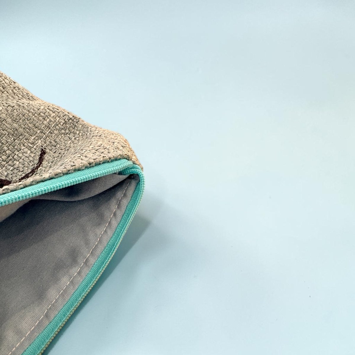 Close-up of a textured beige fabric pouch with a turquoise zipper and gray interior lining, shown on a light blue background.