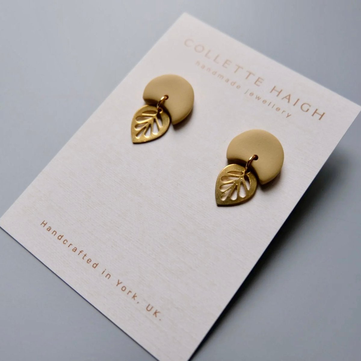 A pair of gold leaf stud earrings mounted on a white display card. The card is branded &