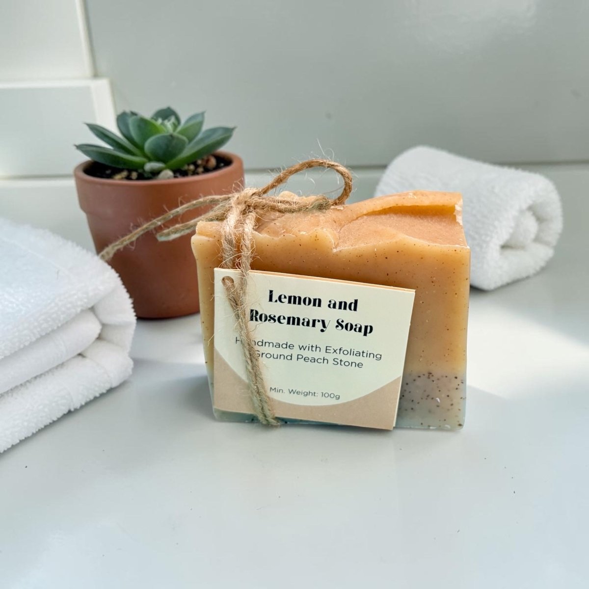 Bar of lemon and rosemary soap with a label on a white surface, surrounded by towels and a potted plant.