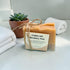 Bar of lemon and rosemary soap with a label on a white surface, surrounded by towels and a potted plant.