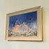 A framed landscape painting of a dramatic blue sky with pink and white clouds over a green and beige field, housed in a light natural wood frame and mounted on a beige wall.