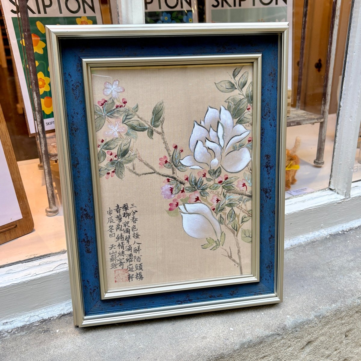 A framed Japanese-style floral artwork leaning against a storefront window. The piece shows large white blossoms, green leaves, and pink accents on a beige background, surrounded by a distressed blue inner frame and a light wood outer frame.
