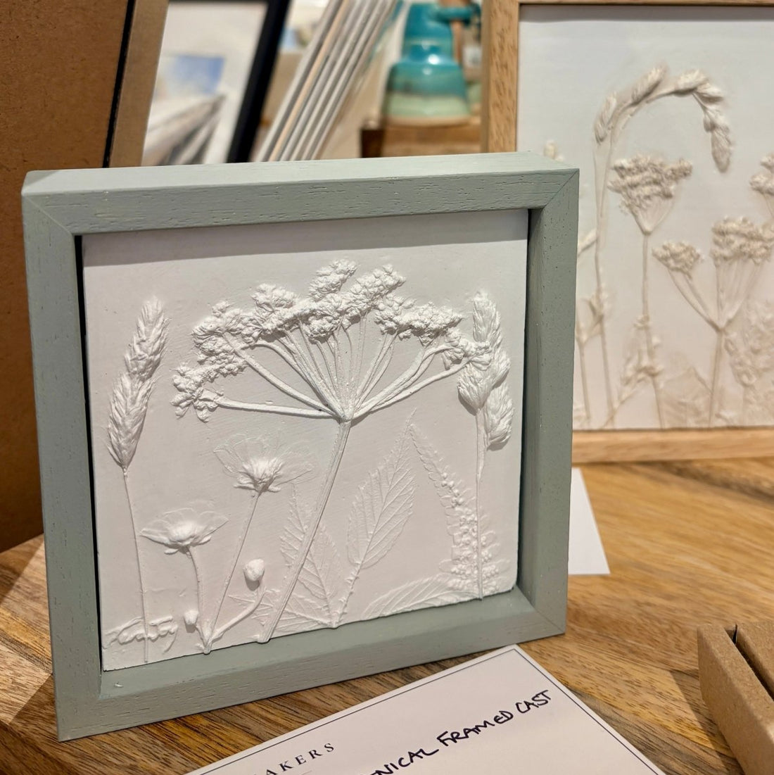 A framed botanical relief artwork showing white raised flower and leaf designs inside a pale sage green frame, displayed on a wooden surface with another similar framed piece in the background.