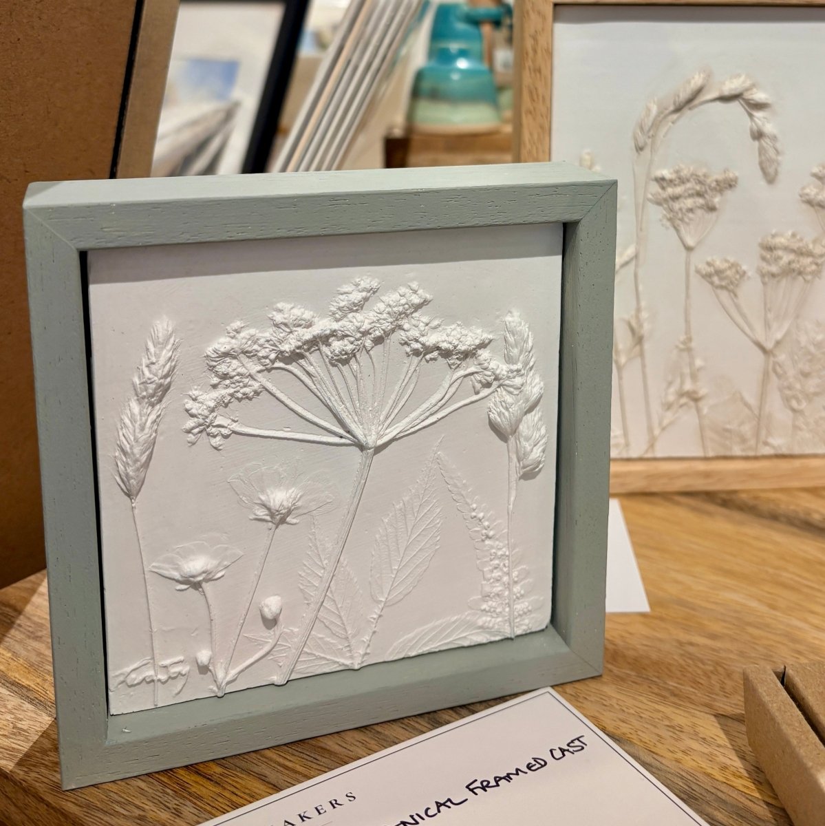A framed botanical relief artwork showing white raised flower and leaf designs inside a pale sage green frame, displayed on a wooden surface with another similar framed piece in the background.
