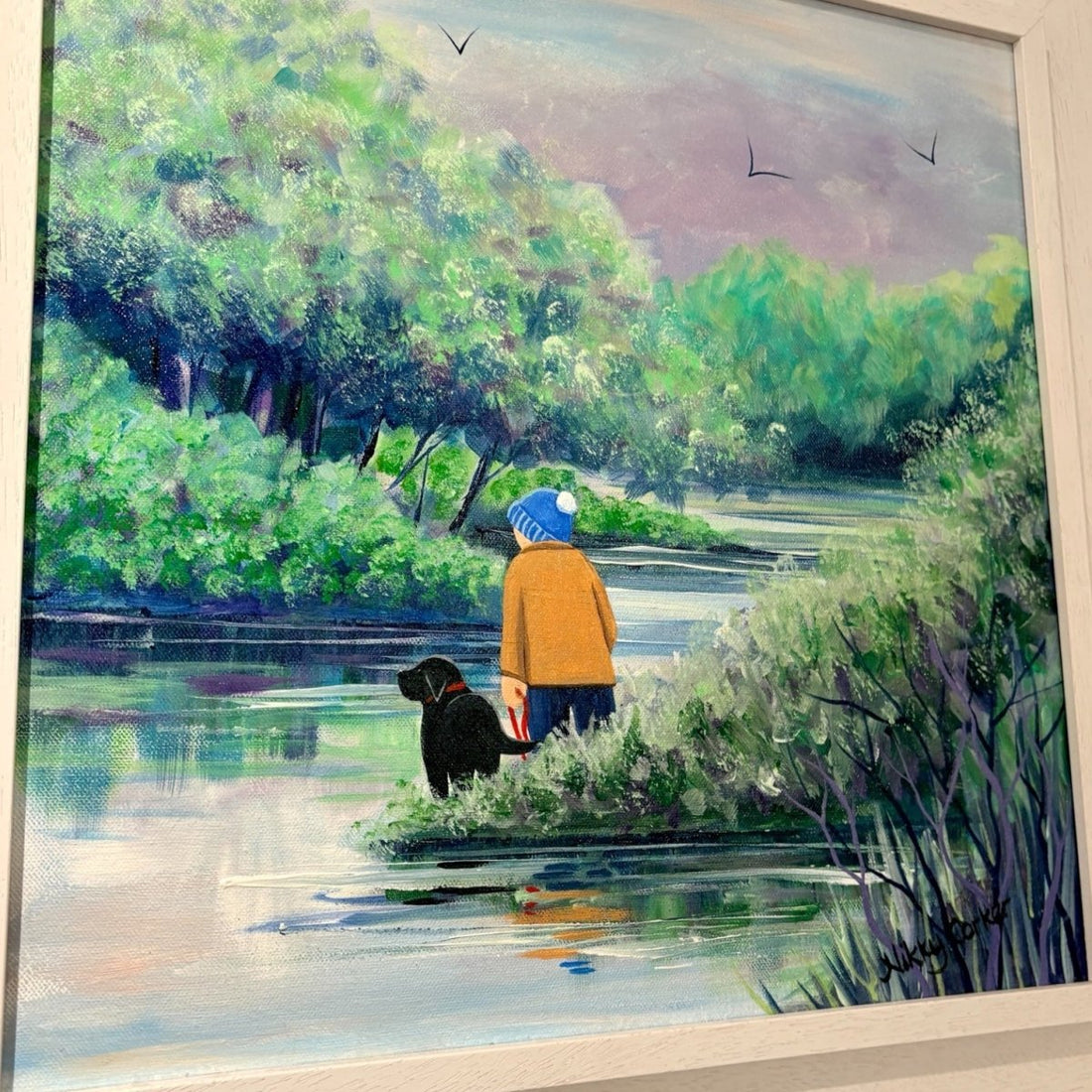 A framed painting depicting a person in a yellow jacket and blue hat standing by a river with a black dog, surrounded by lush green trees and reeds under a soft sky.