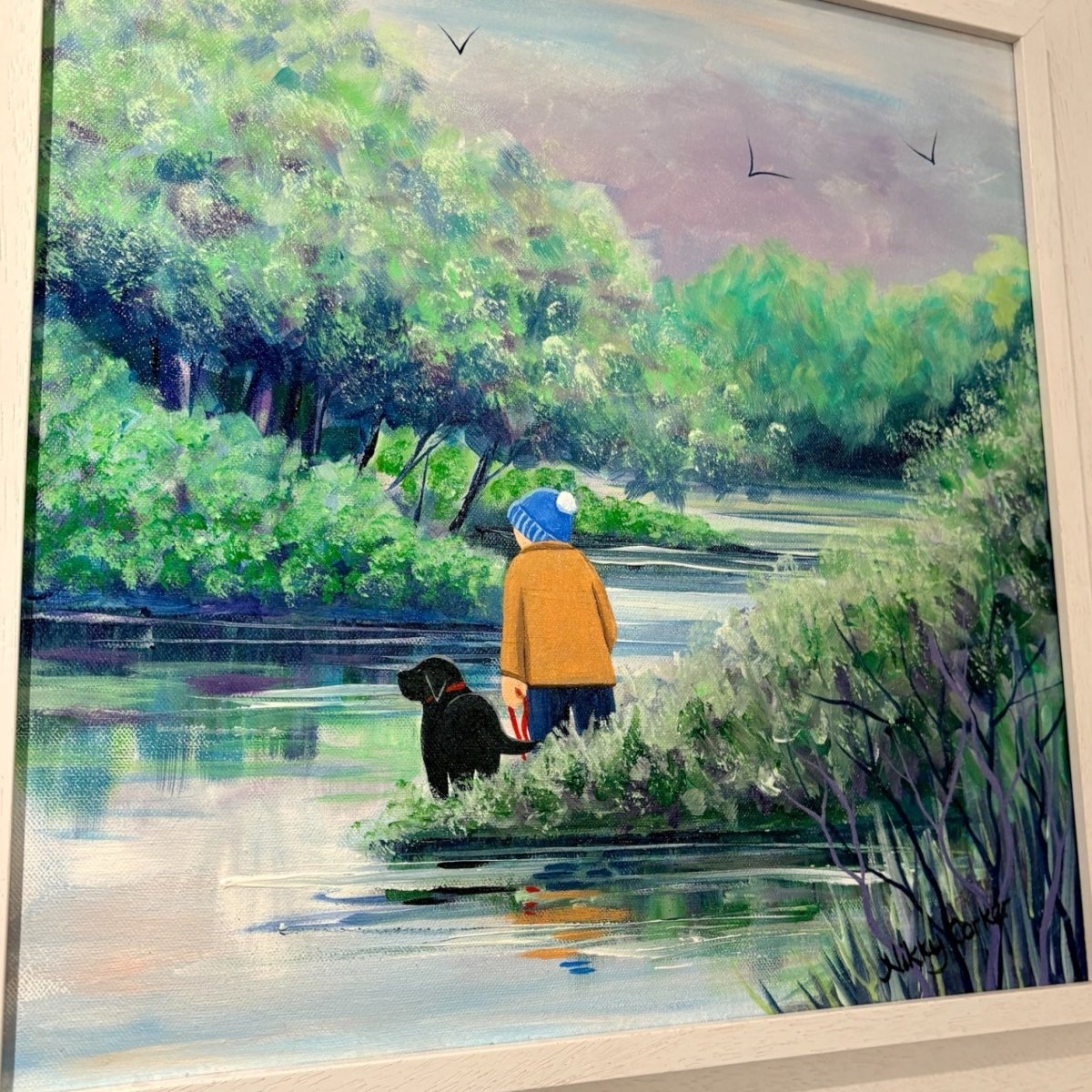 A framed painting depicting a person in a yellow jacket and blue hat standing by a river with a black dog, surrounded by lush green trees and reeds under a soft sky.