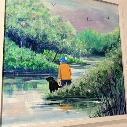 A framed painting depicting a person in a yellow jacket and blue hat standing by a river with a black dog, surrounded by lush green trees and reeds under a soft sky.