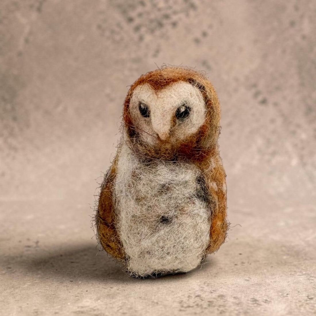 A small felted wool owl figurine with brown and white feathers, standing against a textured beige background.