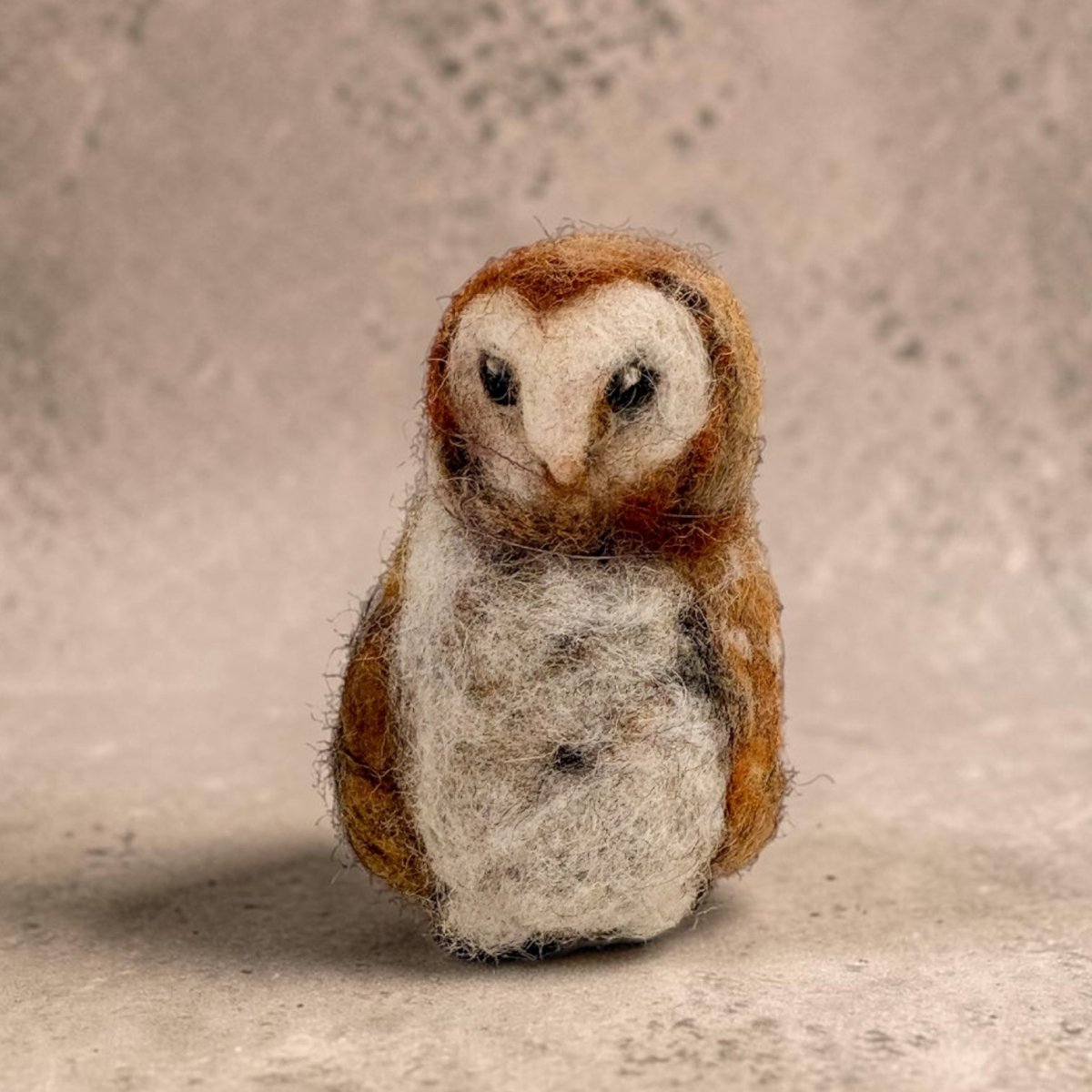 A small felted wool owl figurine with brown and white feathers, standing against a textured beige background.