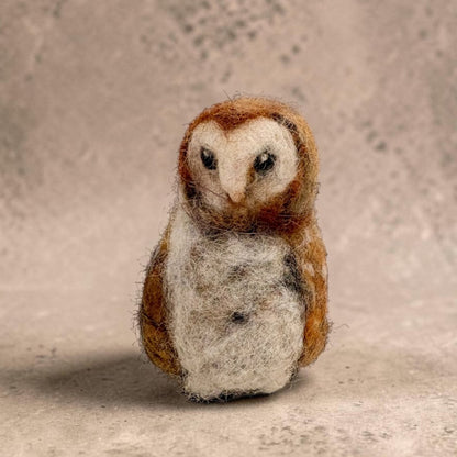 A small felted wool owl figurine with brown and white feathers, standing against a textured beige background.