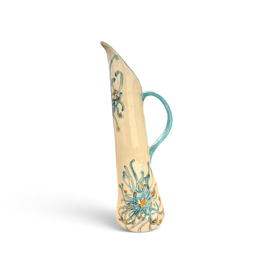 A tall, slender ceramic pitcher with a curved handle, featuring a cream-colored glaze decorated with turquoise blue floral and scroll patterns and small orange accents, set against a plain white background.