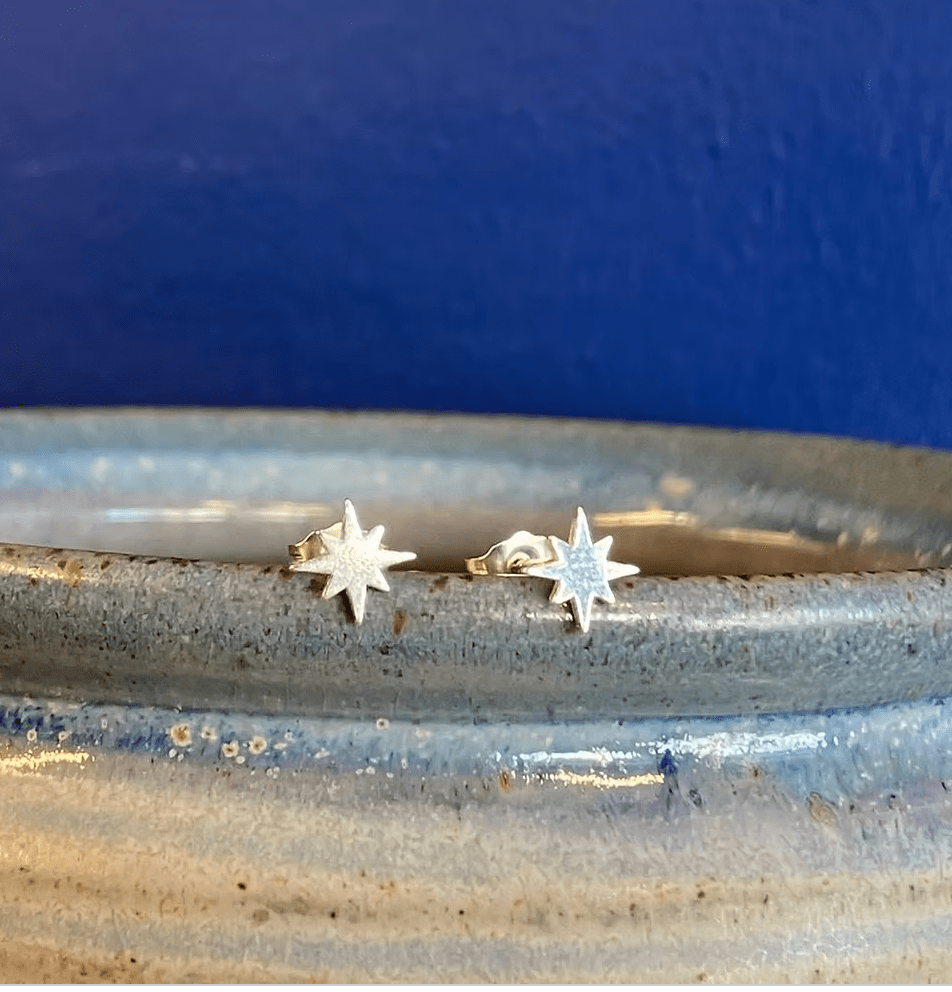 A pair of small silver starburst stud earrings resting on the rim of a speckled ceramic dish against a dark blue background.
