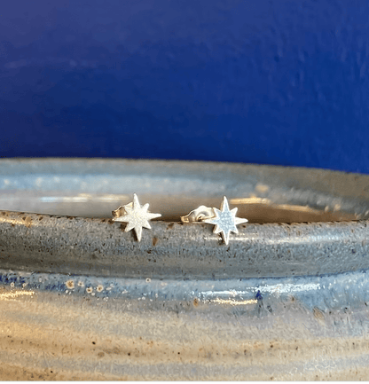 A pair of small silver starburst stud earrings resting on the rim of a speckled ceramic dish against a dark blue background.