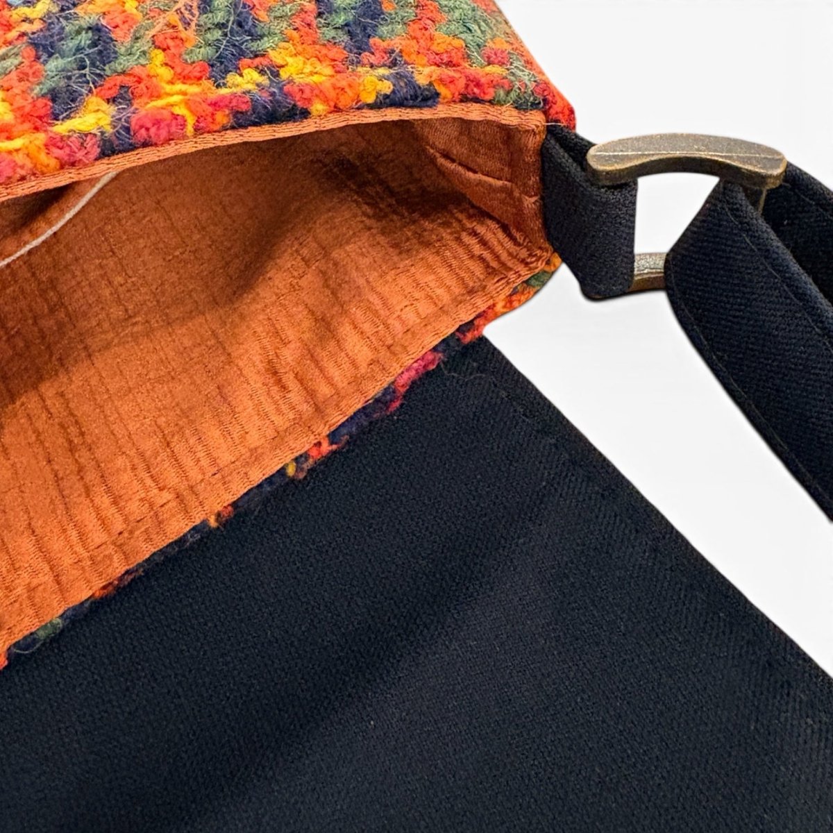 Close-up of a shoulder bag showing a black woven strap with a metal buckle, a dark navy body, and a multicolor textured fabric trim with an orange interior lining.