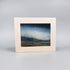 A framed landscape art print with a wide cream-colored border and a textured painting depicting a moody blue sky over a low, dark coastal scene, set against a neutral gray background.