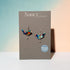 Nancy Jones bird-shaped earrings with a colorful mosaic pattern, displayed on a gray brand card against a pastel blue and peach background.