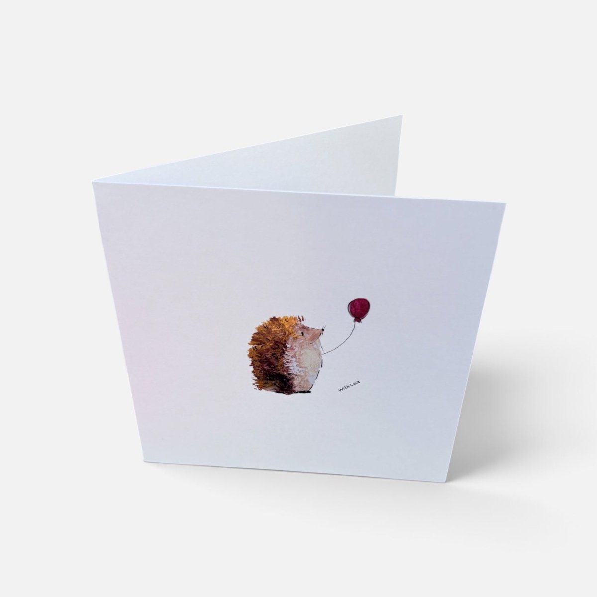 A folded white greeting card standing open on a light gray background, featuring a small watercolor illustration of a hedgehog holding a red balloon.