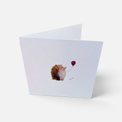 A folded white greeting card standing open on a light gray background, featuring a small watercolor illustration of a hedgehog holding a red balloon.