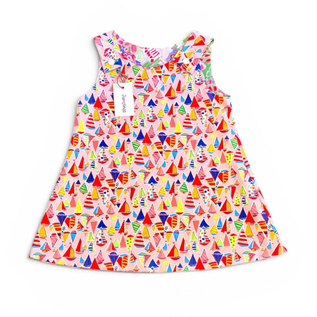 A sleeveless baby dress with a colorful sailboat print on a light pink background, featuring a ruffled neckline and a white hangtag.
