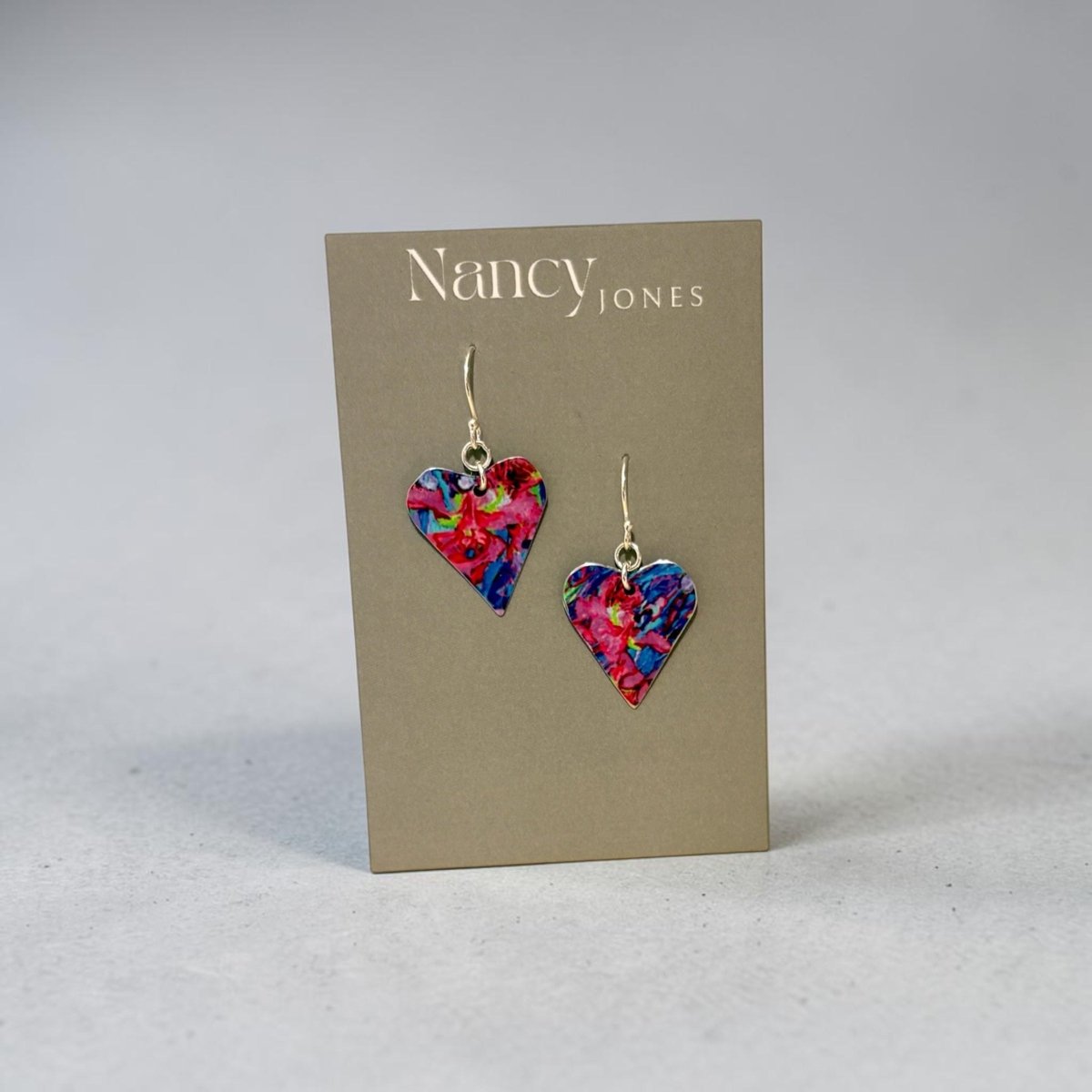 A pair of heart-shaped dangling earrings with a vibrant multicolor floral pattern, displayed on a brown Nancy Jones brand card against a neutral gray background.