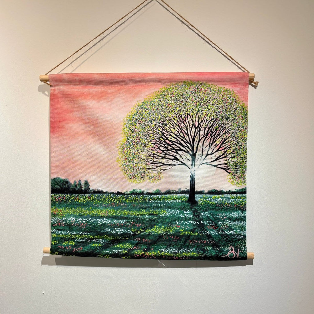 A square fabric wall hanging scroll with a wooden dowel and hanging cord, featuring a painted landscape of a large tree in a field under a pink sky, displayed on a light wall.