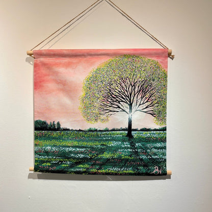 A square fabric wall hanging scroll with a wooden dowel and hanging cord, featuring a painted landscape of a large tree in a field under a pink sky, displayed on a light wall.