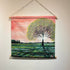 A square fabric wall hanging scroll with a wooden dowel and hanging cord, featuring a painted landscape of a large tree in a field under a pink sky, displayed on a light wall.
