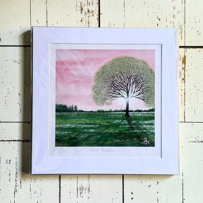 Framed artwork of a tree in a field with a pink sky on a wooden surface