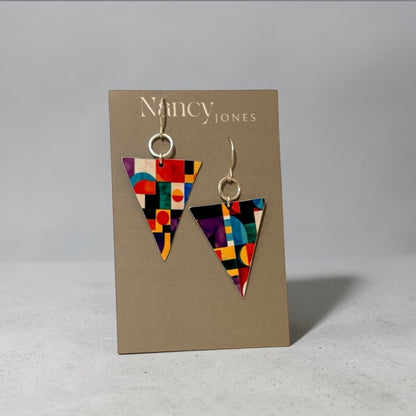 Triangle dangle earrings with a colorful geometric abstract pattern, displayed on a brown Nancy Jones brand card against a neutral gray background.