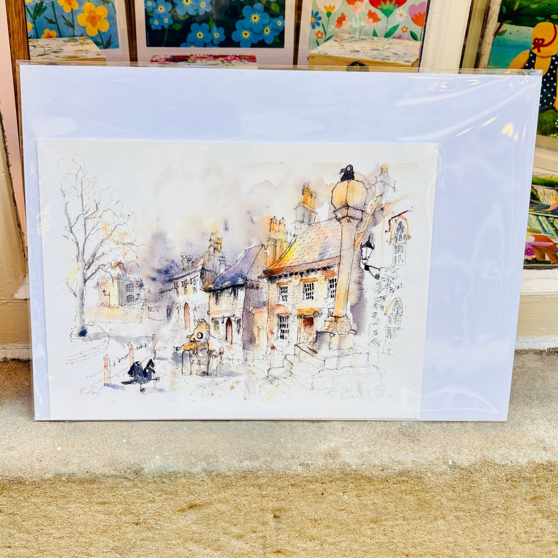 A watercolor art print depicting a quaint village street with old stone buildings, a tree, and a streetlamp, shown in its clear plastic protective sleeve on a shelf.