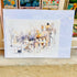 A watercolor art print depicting a quaint village street with old stone buildings, a tree, and a streetlamp, shown in its clear plastic protective sleeve on a shelf.