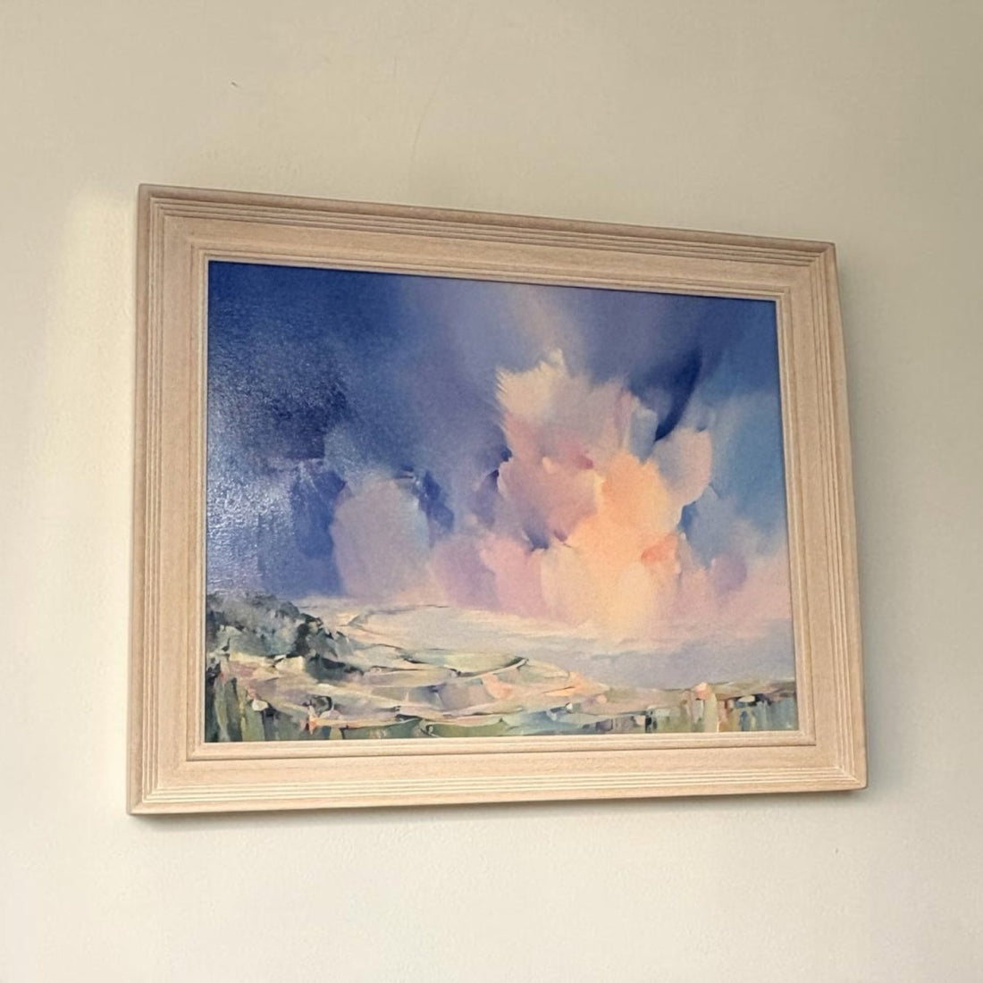 A framed landscape painting hanging on a light beige wall. The artwork depicts dramatic blue and pink clouds over a pale landscape, enclosed in a light natural wood frame.