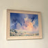 A framed landscape painting hanging on a light beige wall. The artwork depicts dramatic blue and pink clouds over a pale landscape, enclosed in a light natural wood frame.