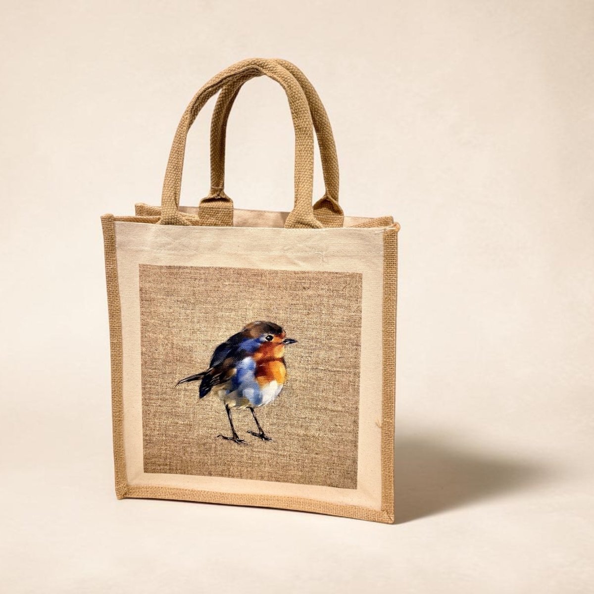 A natural jute tote bag with a central printed panel featuring a bluebird on a burlap-textured background, set against a warm beige studio backdrop.