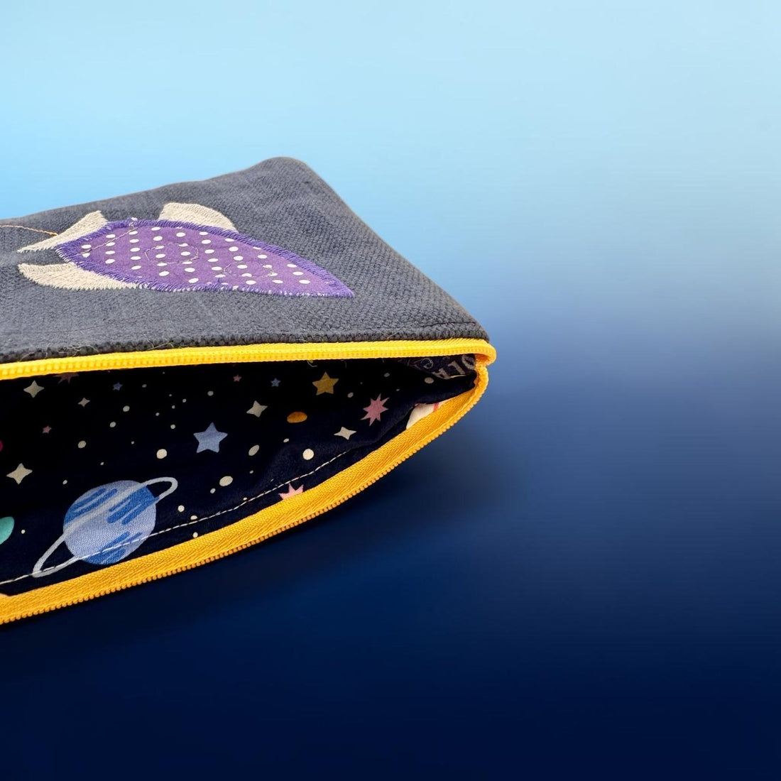 A fabric zippered pouch with a dark gray exterior featuring a purple rocket illustration, and a black space-themed interior printed with planets and stars, finished with a bright yellow zipper trim.