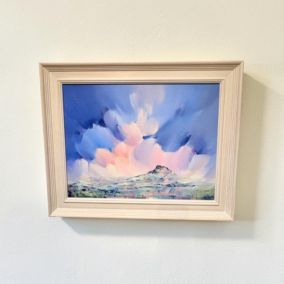 A framed landscape painting featuring a dramatic blue and pink sky with white clouds and a small mountain, displayed on a light beige wall.