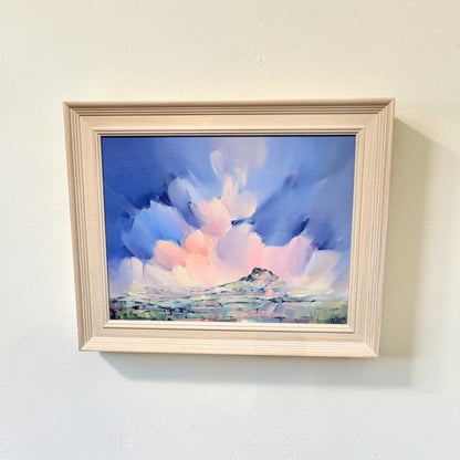 A framed landscape painting featuring a dramatic blue and pink sky with white clouds and a small mountain, displayed on a light beige wall.